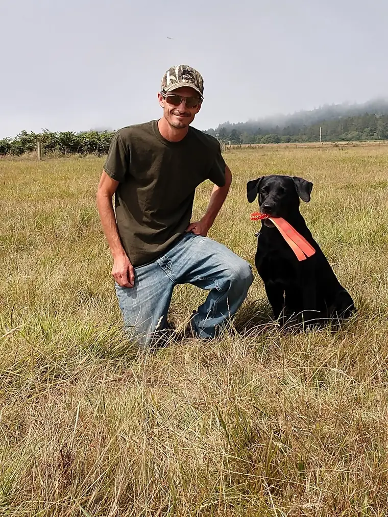 Richard Gebhart kneeling in a grass field beside a black Labrador holding an orange ribbon